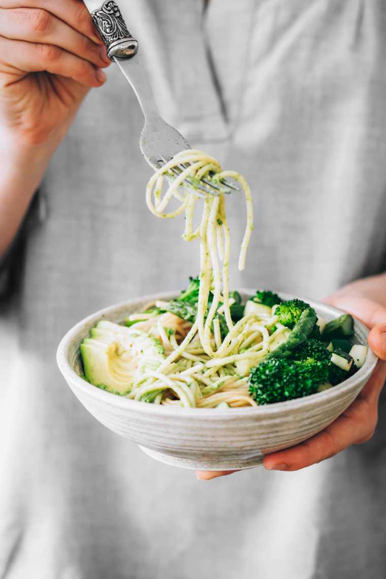 woman in grey shirt holding a bowl of pasta with green veggies and sauce