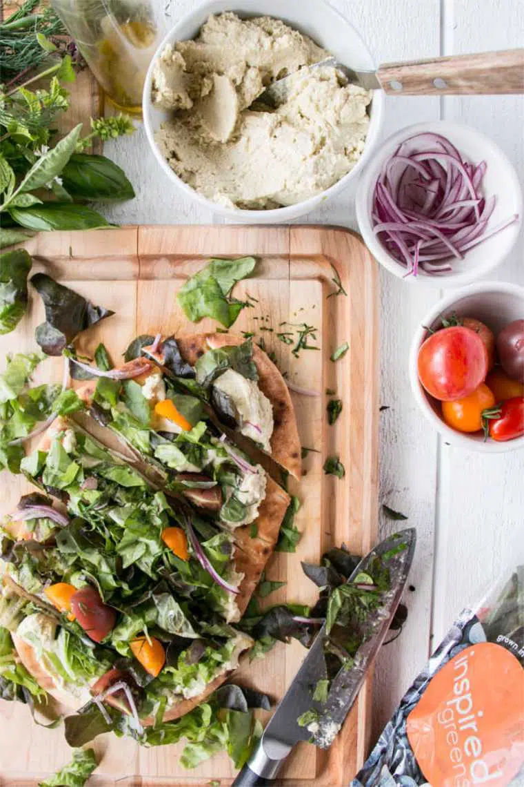 wooden chopping board with a flatbread that's spread with hummus, topped with veggies and cut into pieces