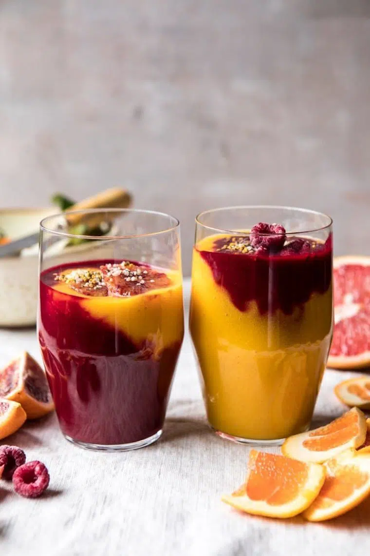 two glasses of bright orange and dark red fruity citrus smoothie next to some fruit and a knife