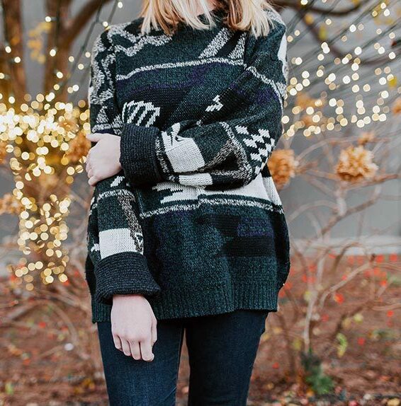 woman in black knitted sweater with white patterns standing in front of an autumn tree full of small lights during the holidays