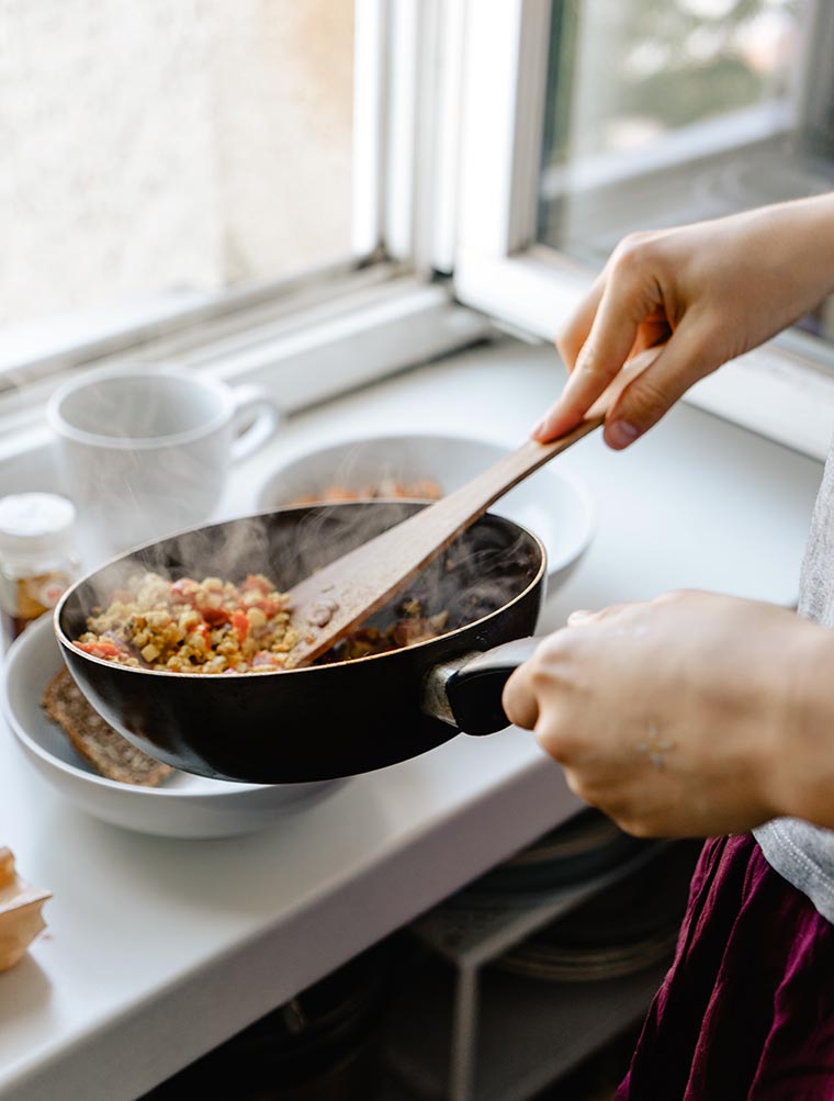 person holding a black pan with cooked lentils and vegetables in her hand and standing next to an open window