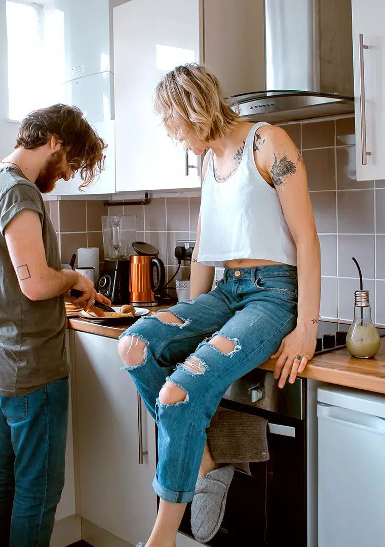 Man cutting vegetables in the kitchen next to a Woman in jeans sitting on the counter