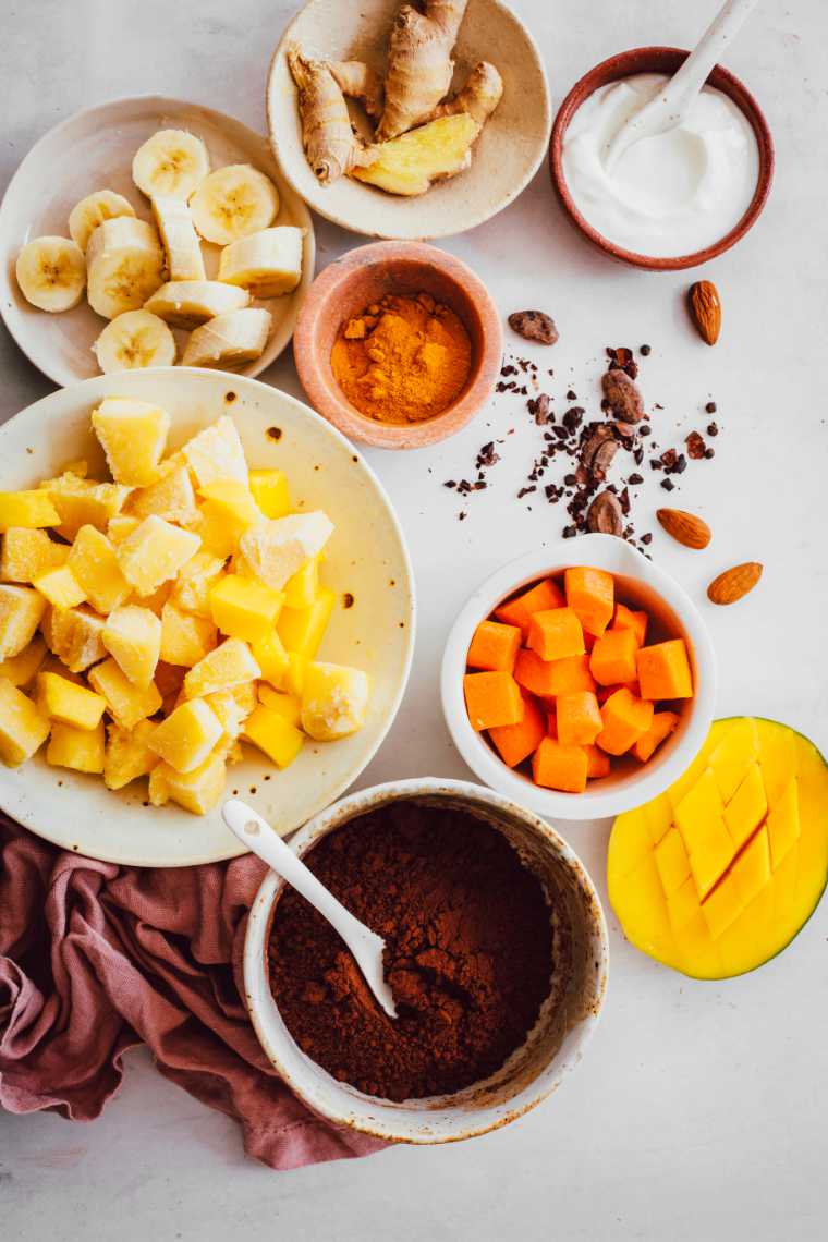 white table with almonds, mango and several bowls with cocoa powder, banana, turmeric and ginger