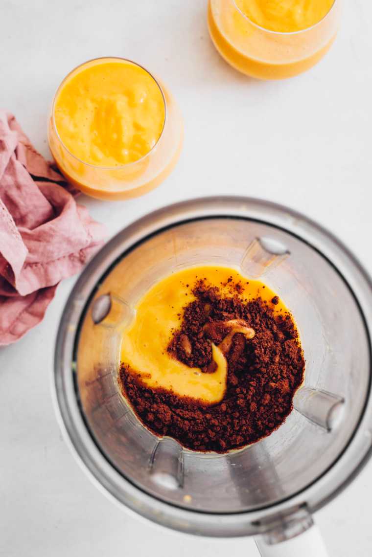 blender jar with mango smoothie and cocoa powder next to glasses of mango smoothie