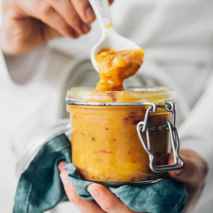 woman in apron holding a glass jar with homemade yellow Indian sugar-free mango chutney on a spoon