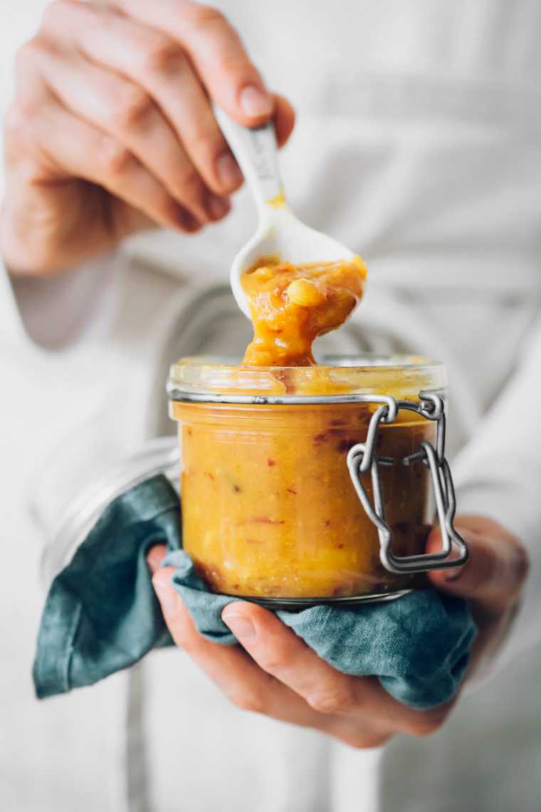 woman in apron holding a glass jar with homemade yellow Indian sugar-free Indian dipping sauce on a spoon