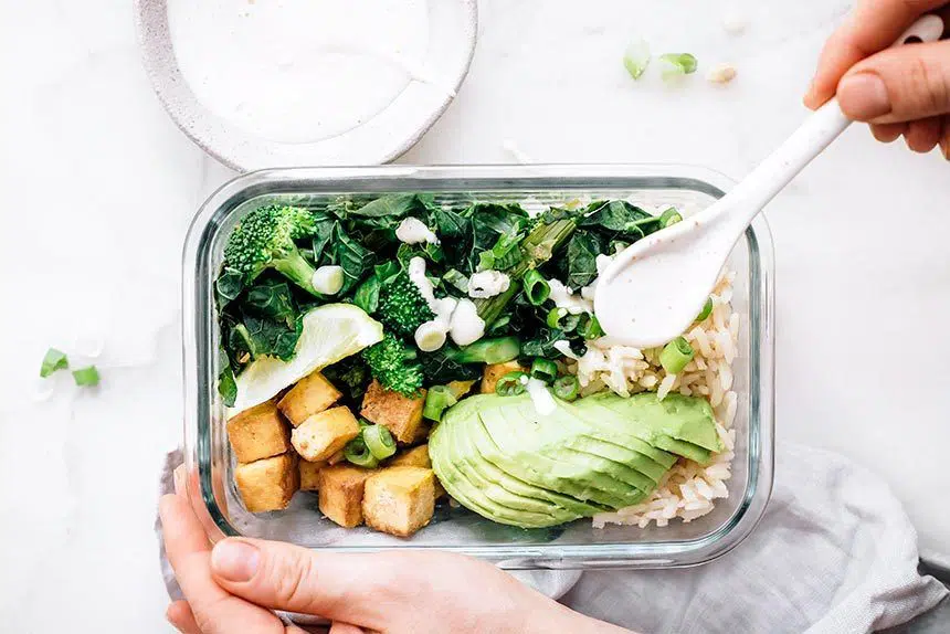 Woman drizzling white sauce over glass bowl filled with rice, avocado, broccoli, green onion and tofu for plant-based meal prep