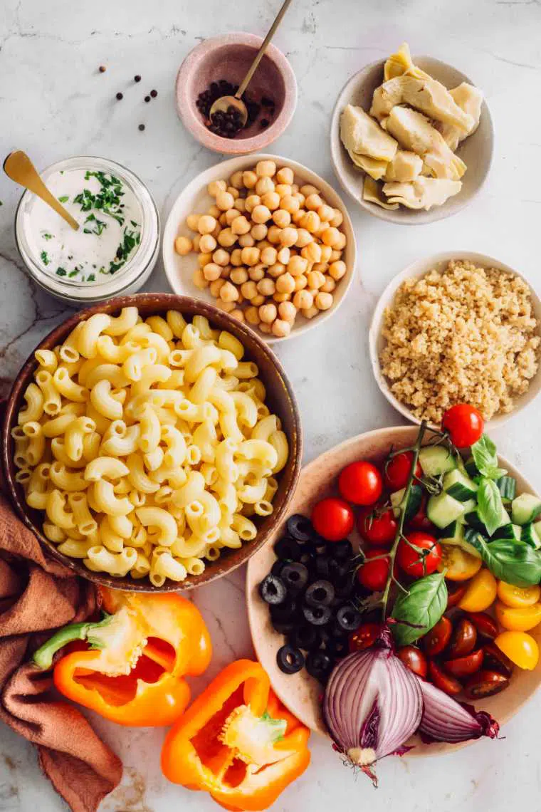 white table with different sized bowls containing cooked quinoa, chickpeas, artichokes, macaroni, bell pepper and salad veggies