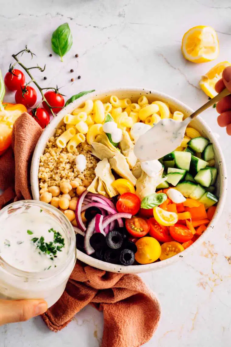 large white bowl with pasta, quinoa, chickpeas and chopped fresh veggies which is being topped with vegan Greek yogurt dressing