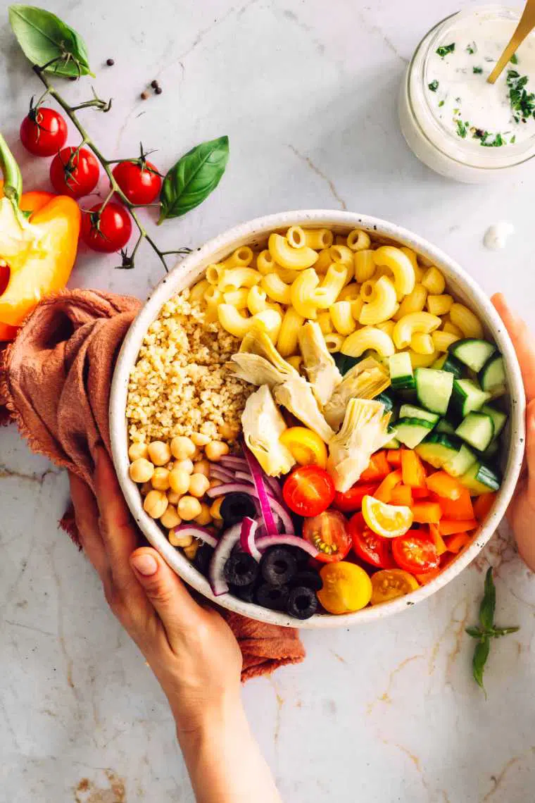 woman holding a large white bowl with quinoa, chickpeas, olives, pasta, cucumber and tomatoes