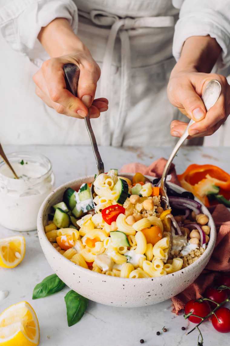 woman in beige clothes standing by a table with a large white bowl containing a vegan Mediterranean quinoa pasta salad and mixing it with a spoon