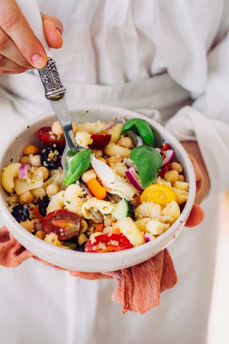woman in white apron holding a bowl of vegan quinoa pasta salad with tomatoes, olives, chickpeas and basil