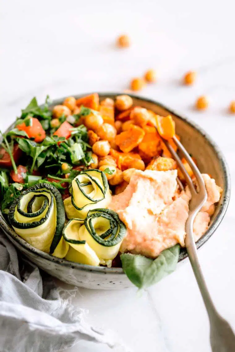 plant based bowl with chopped leafy greens, tomato, sweet potato cubes, chickpeas, zucchini and a dollop of homemade sundried tomato hummus next to a fork