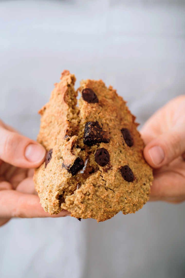 woman wearing a grey top breaking a vegan oatmeal cookie with chocolate chips in half