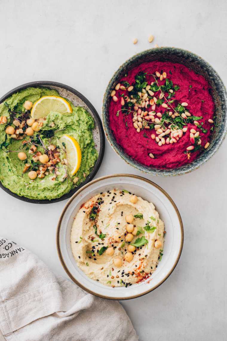 white table with three bowls of different-colored hummus from beige to green spinach and red beet