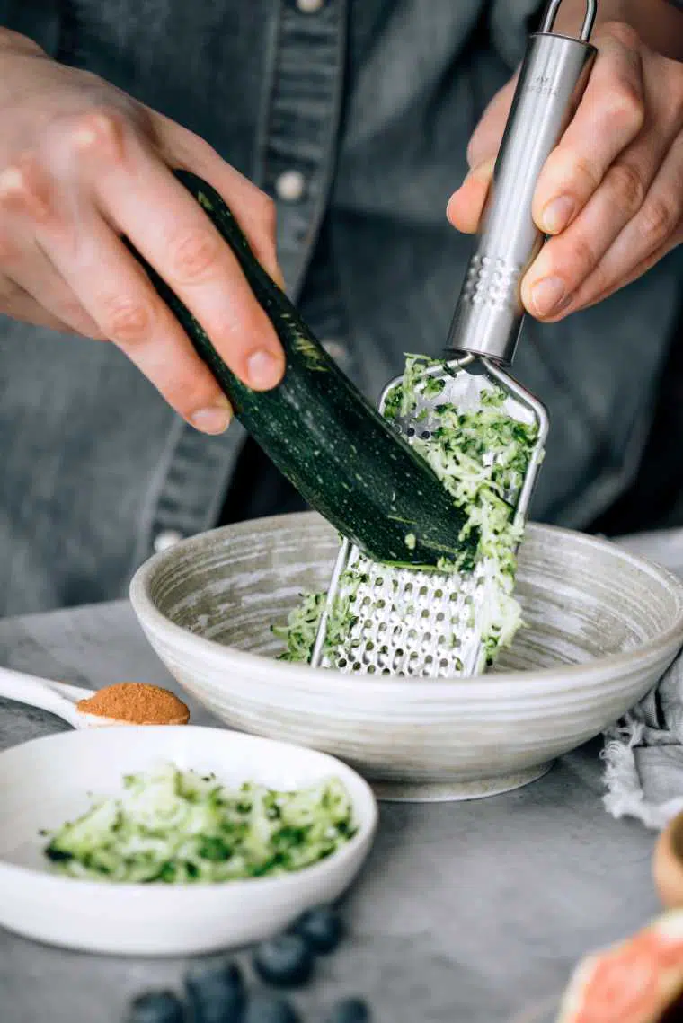 woman in jeans jacket grating some zucchini into a white bowl