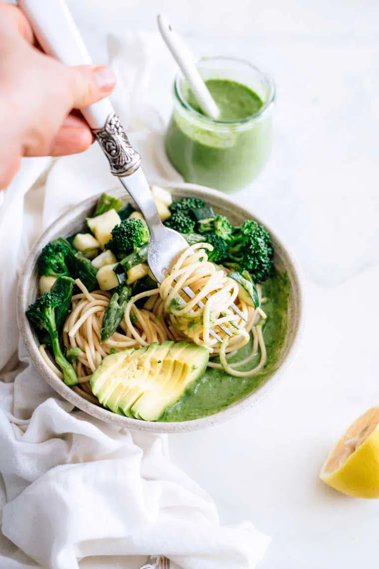 hand with a fork rolling up some soba noodles in a bowl with sliced avocado, broccoli, asparagus and zucchini which is topped with homemade vegan pesto