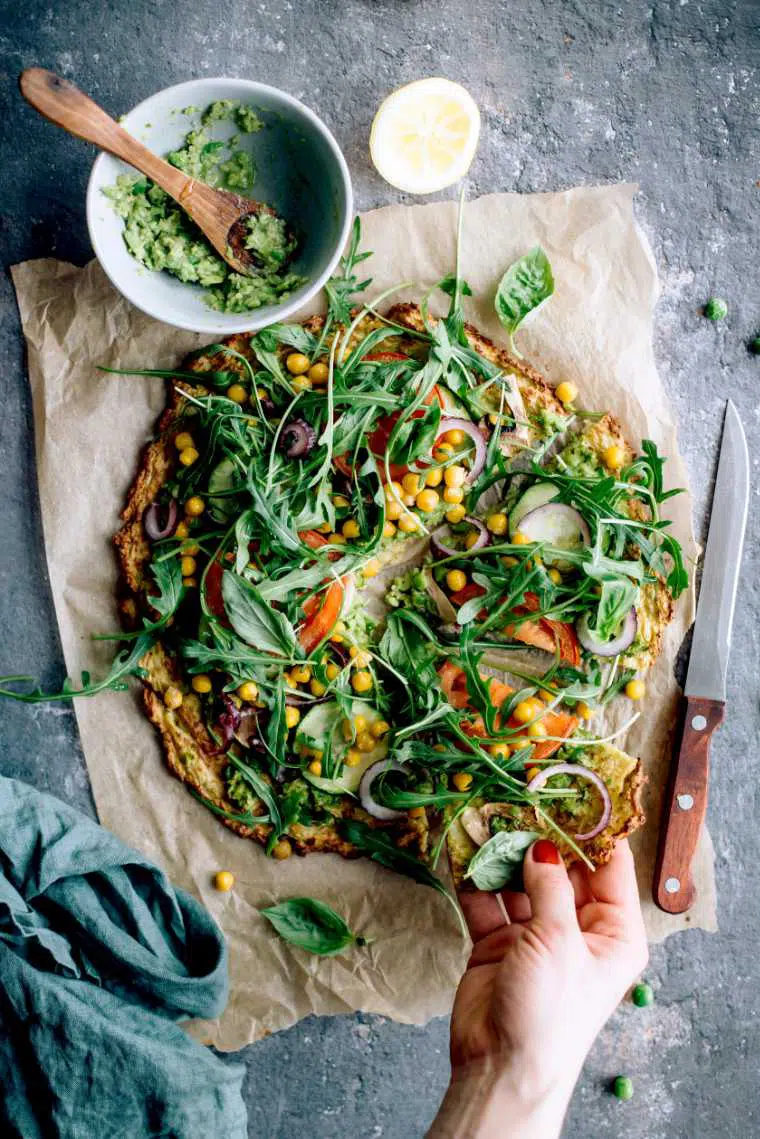 baked vegan cauliflower crust pizza with veggies of which is one slice taken by a hand