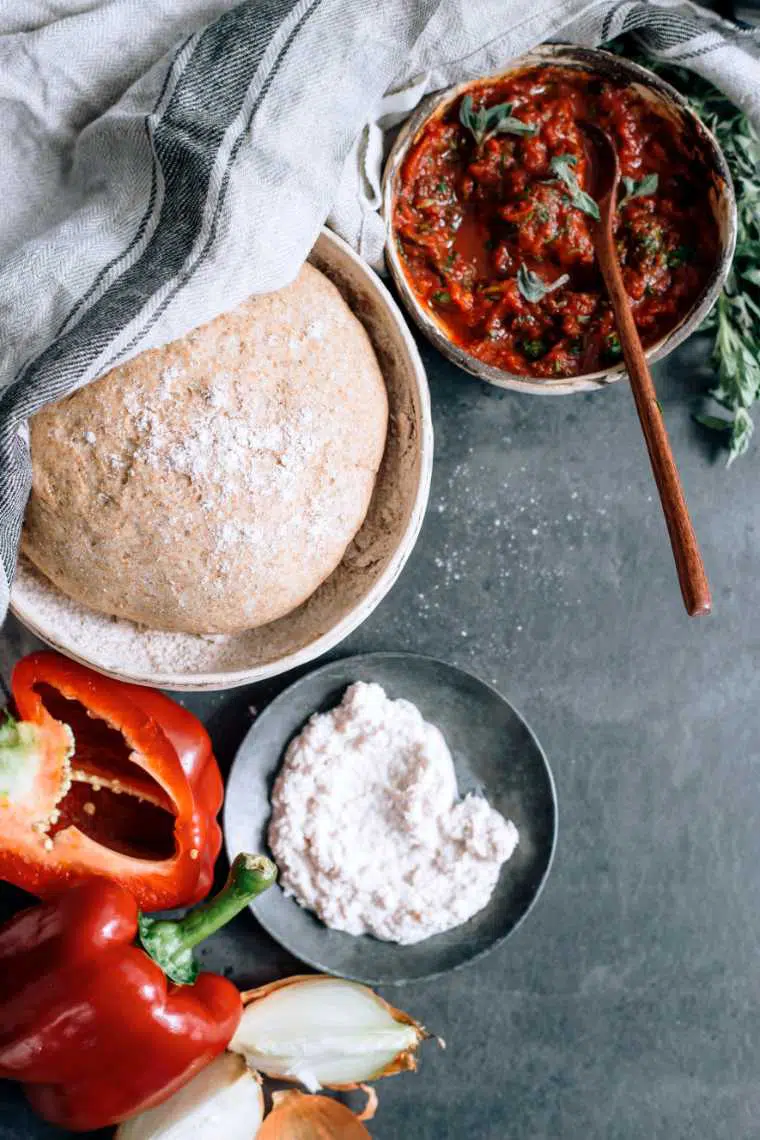 grey table with onions, red bell pepper, tomato sauce and whole wheat dough in a bowl