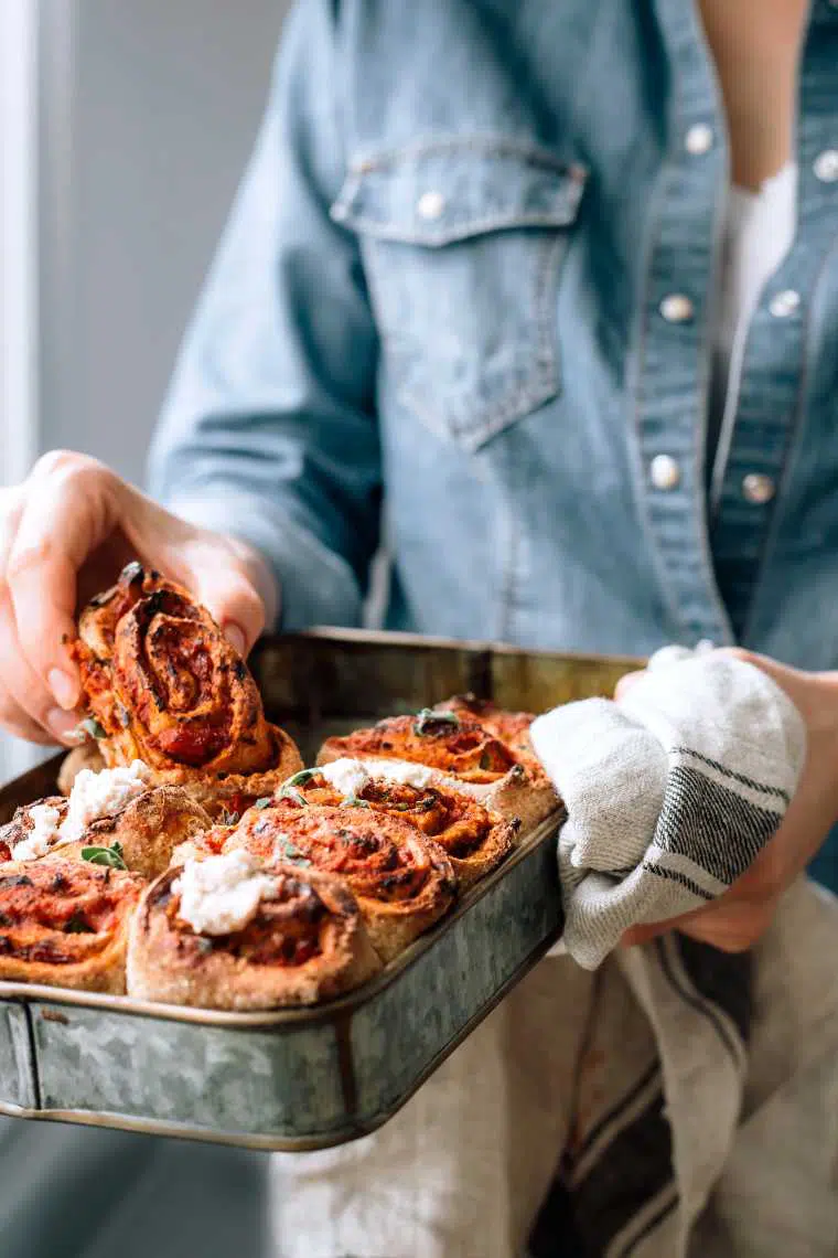 woman in jeans jacket holding a tray of freshly baked healthy vegan pizza rolls with almond ricotta and taking one to eat it