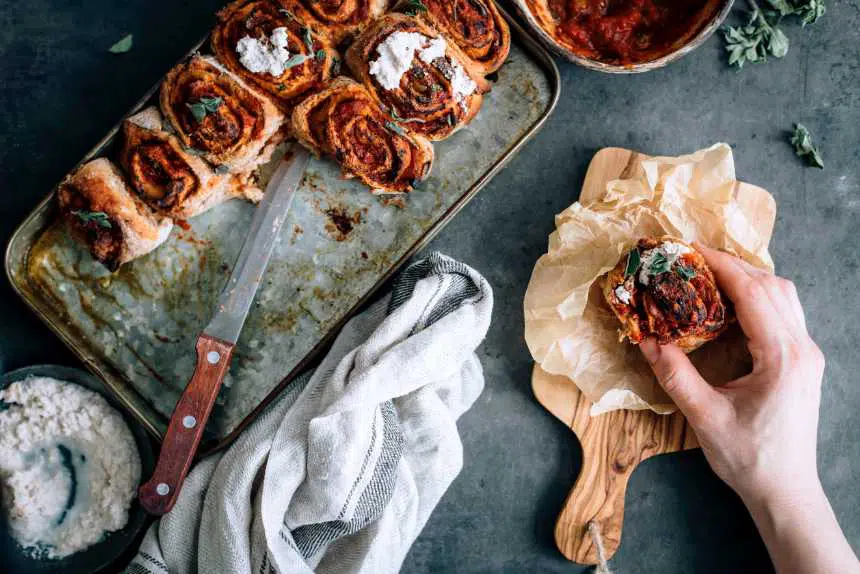 baked vegan pizza pinwheels on a baking dish, one of which is being picked up by a hand from a wooden chopping board