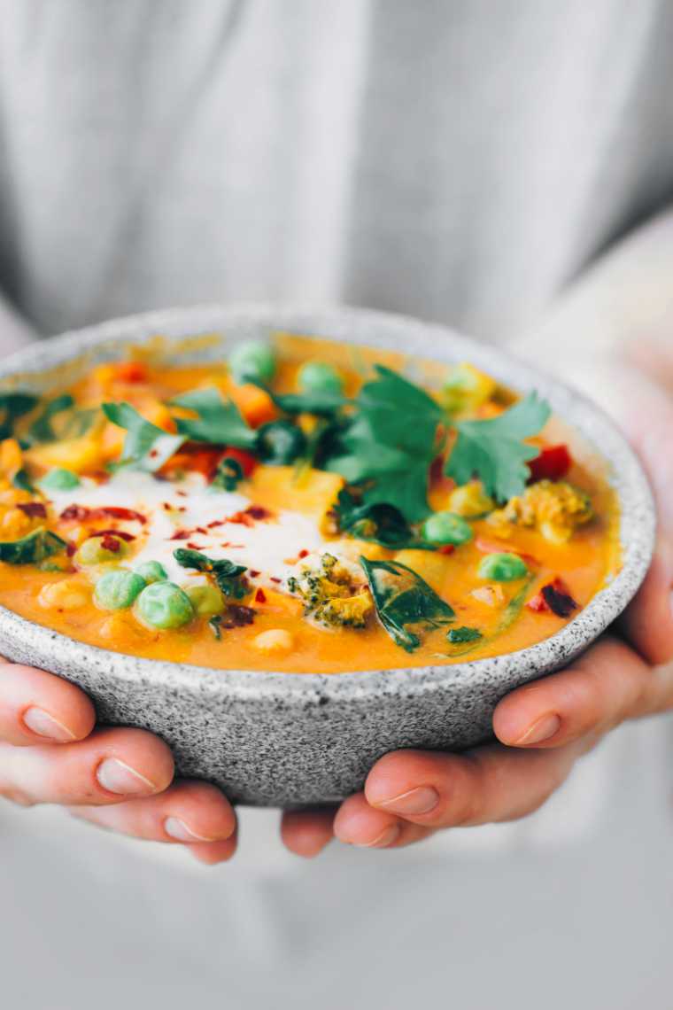 Closeup of woman in grey shirt holding bowl with finished vegan curry in front of her