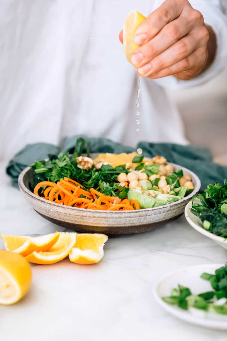woman drizzling lemon juice over a vegan bowl containing spiralized carrot, sliced zucchini, quinoa, chickpeas, oranges and green onion
