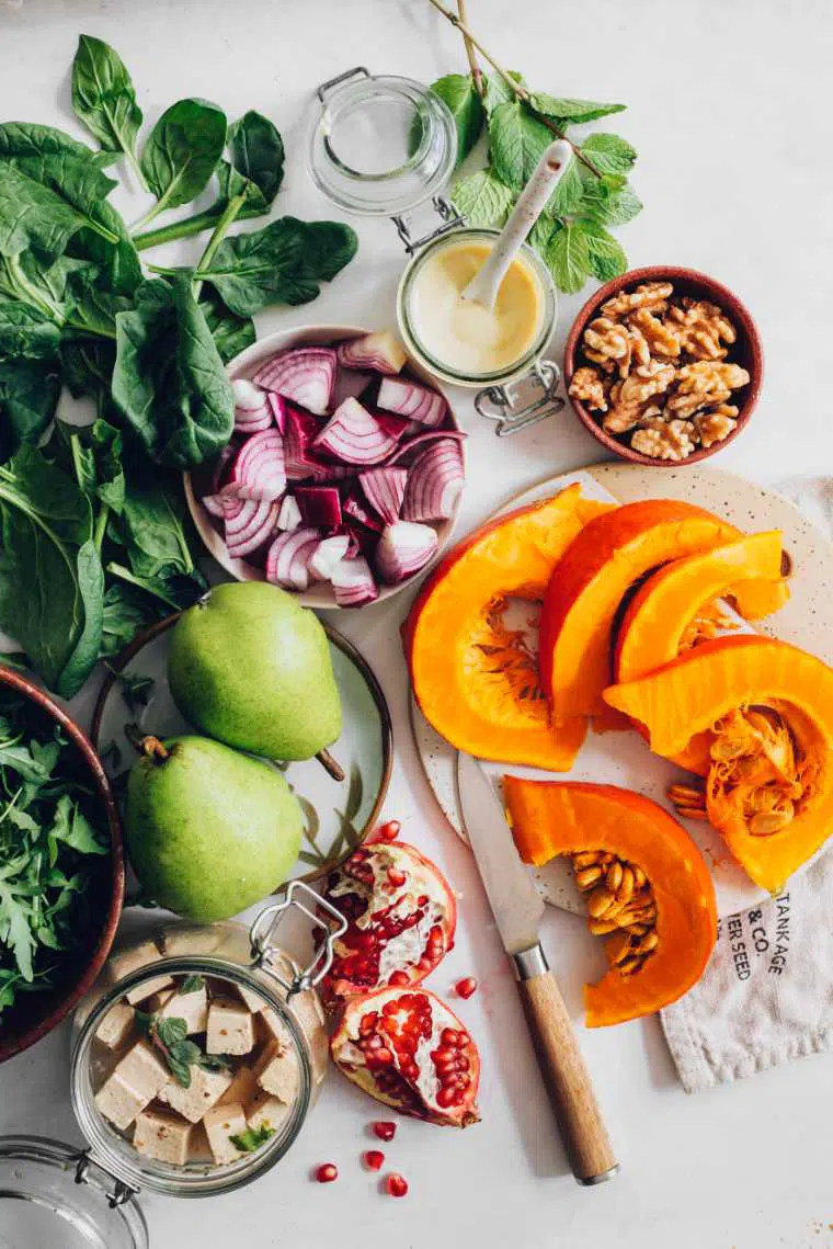 table with sliced pumpkin, red onion, walnuts, pomegranate, pear, spinach and tofu feta