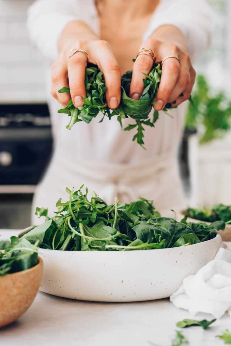 woman in white shirt and apron leaning over a table and holing fresh spinach and arugula in her hands