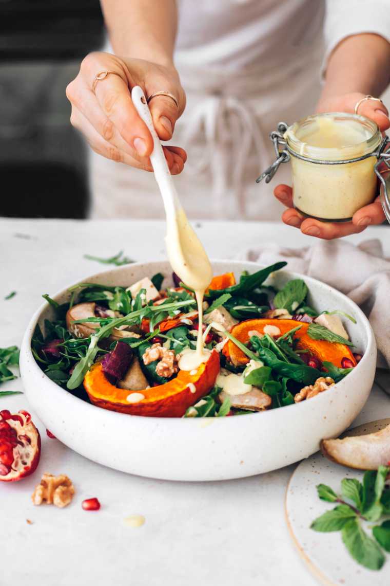 woman in apron drizzling mustard dressing over roast pumpkin and spinach salad in a bowl