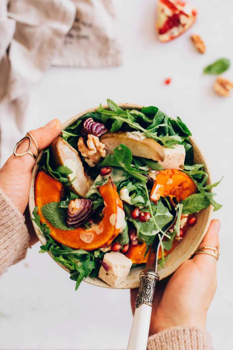 woman in brown sweater holding a small bowl of colorful spinach, pumpkin, feta and pear salad in her hands