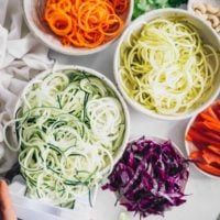 several bowls of spiralized squash, zucchini and carrot as well as sliced red cabbage and bell pepper on a table