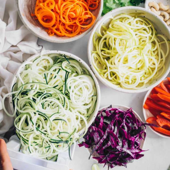 several bowls of spiralized squash, zucchini and carrot as well as sliced red cabbage and bell pepper on a table