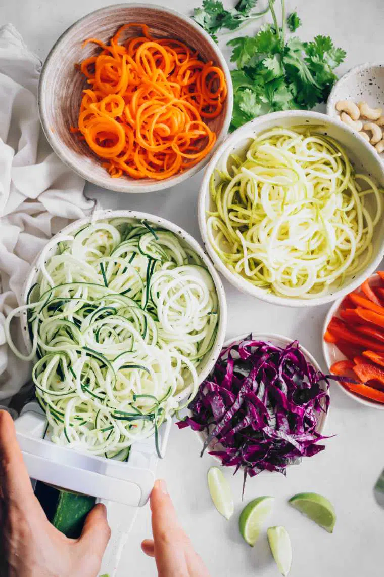 several bowls of spiralized squash, zucchini and carrot as well as sliced red cabbage and bell pepper on a table