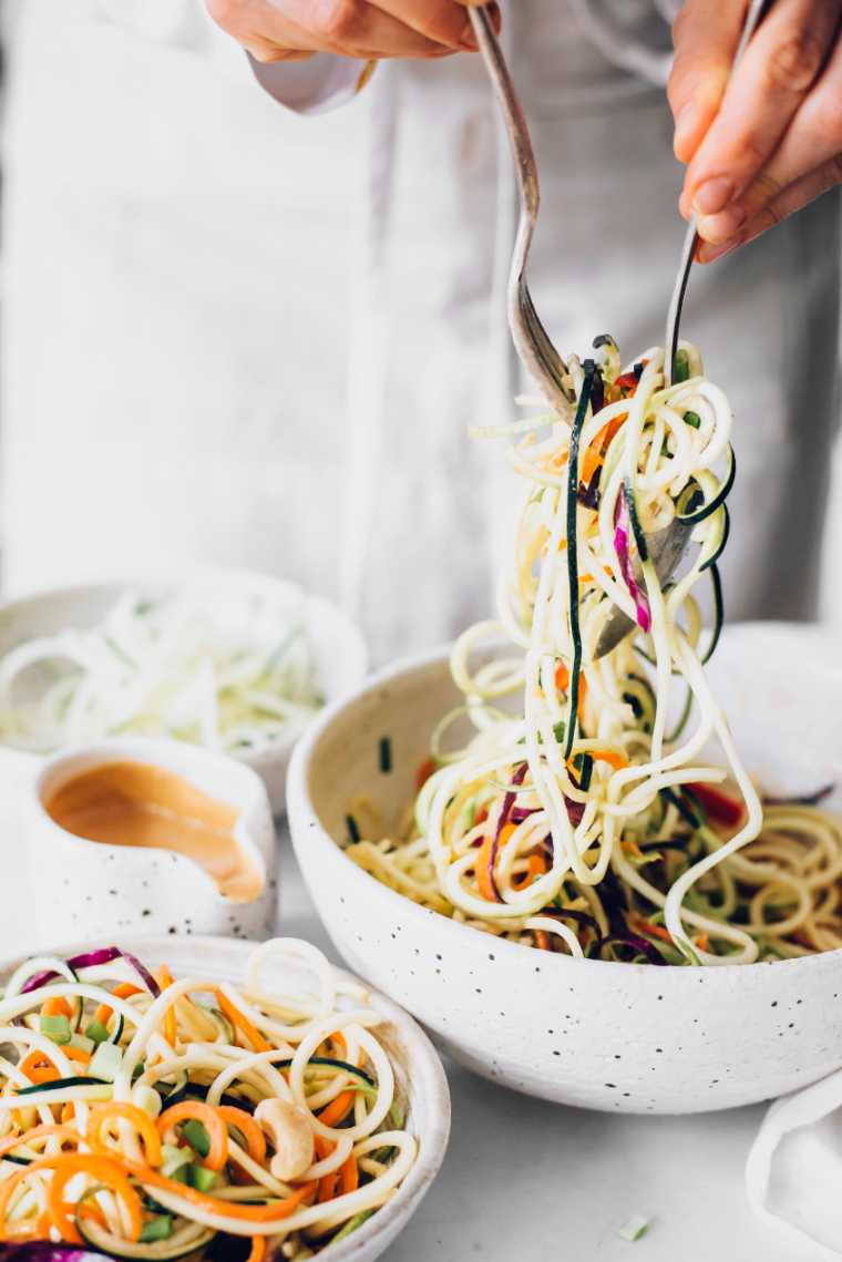 woman in white apron mixing spiralized raw vegetables in a large bowl with a fork and a spoon