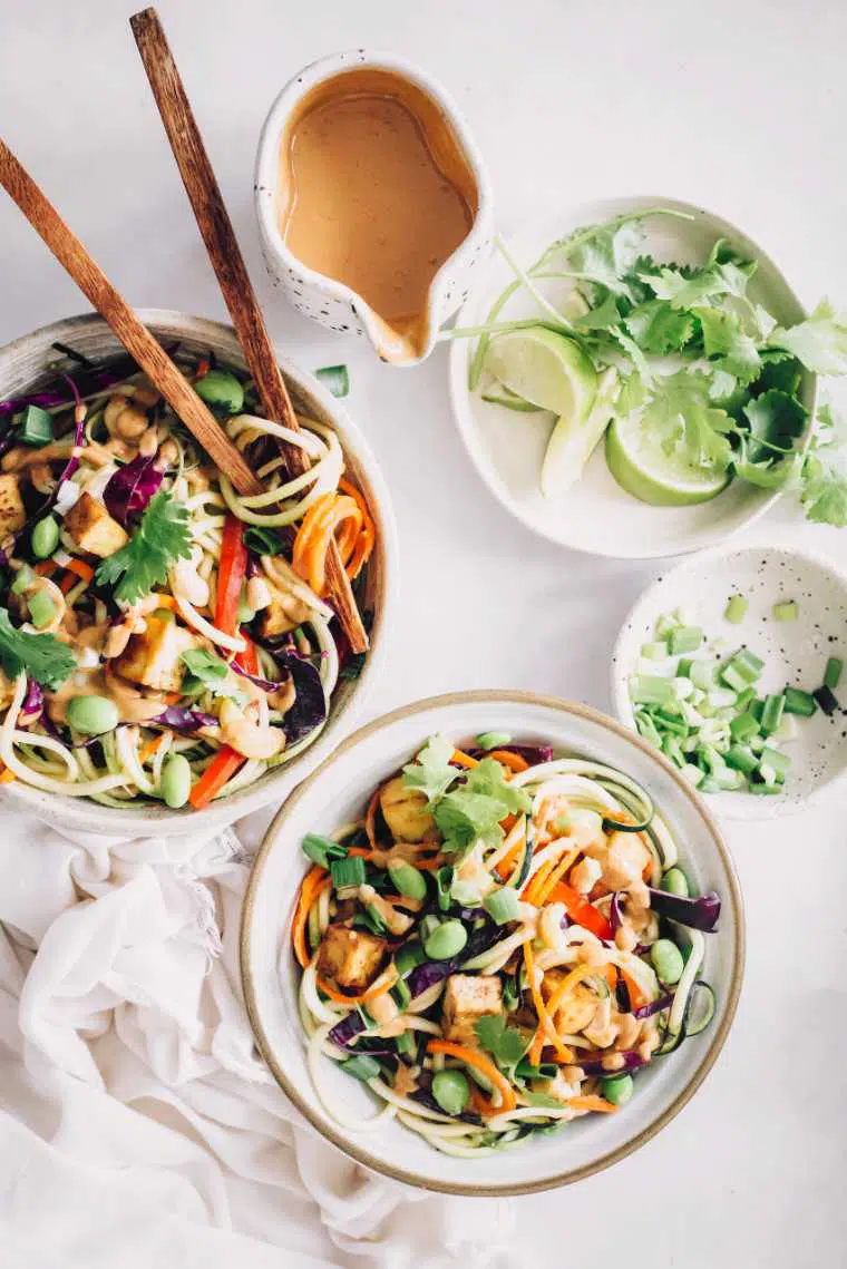 two bowls of colorful spiralized veggies topped with edamame, tofu and a creamy peanut butter sauce