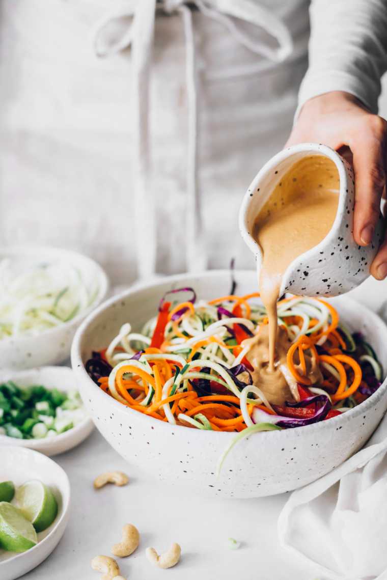 woman in white apron standing in front of a large white bowl with raw vegan pad thai and pouring peanut butter sauce over the vegetables