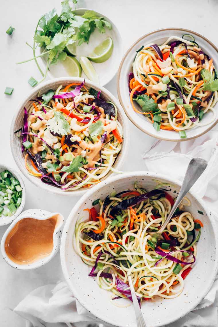 top view of white table with three different-sized bowls containing spiralized zucchini, carrots and more veggies