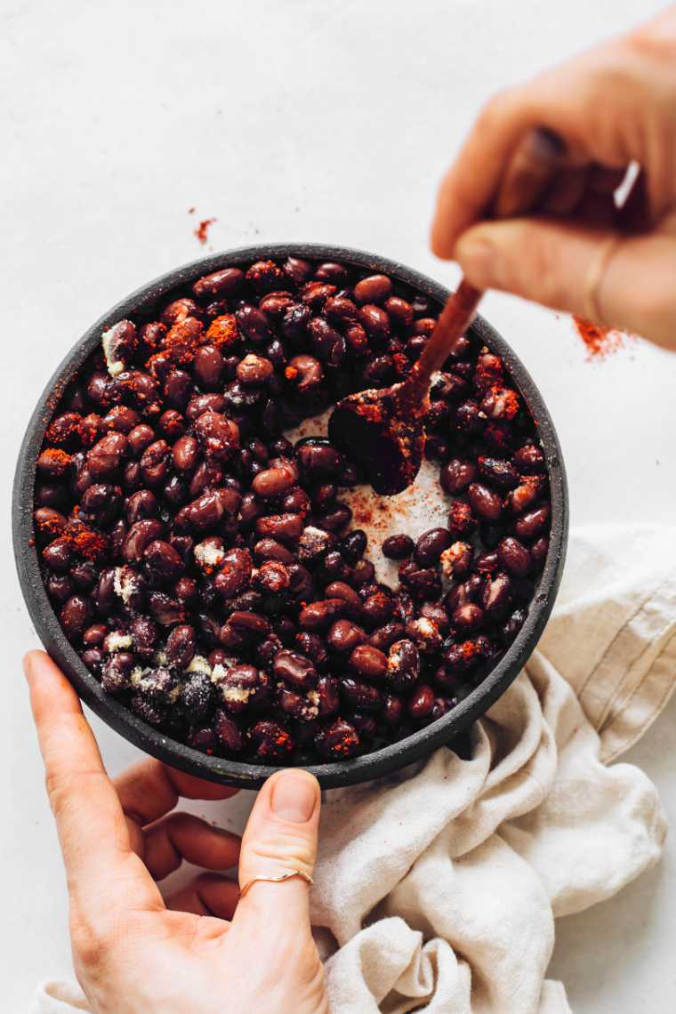 two hands holding a bowl and a spoon while mixing black beans with spices