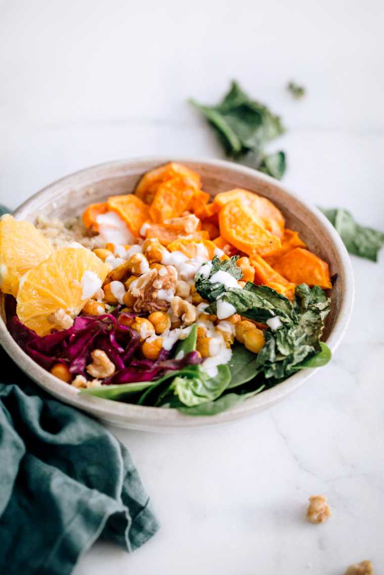 white bowl on a bright table with colorful veggies, such as spinach, red cabbage, sweet potatoes and some quinoa, chickpeas and sliced orange drizzled with a white vegan dressing