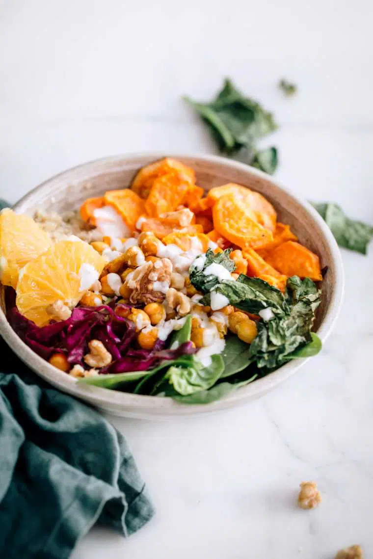 white bowl on a bright table with colorful veggies, such as spinach, red cabbage, sweet potatoes and some quinoa, chickpeas and sliced orange drizzled with a white vegan dressing
