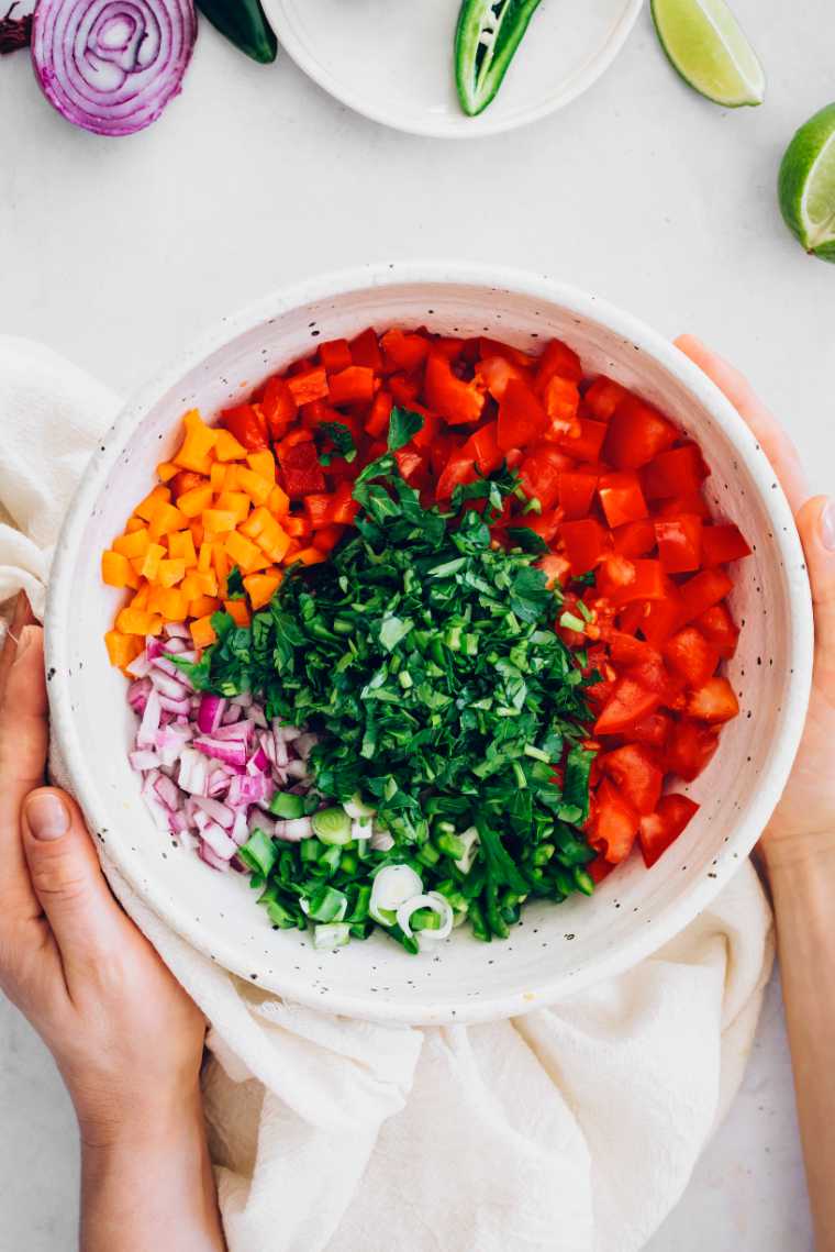 woman holding large white bowl containing chopped herbs, tomatoes, onion and carrots to make sugar free salsa at home