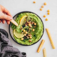 hand holding a breadstick and dipping it into green hummus