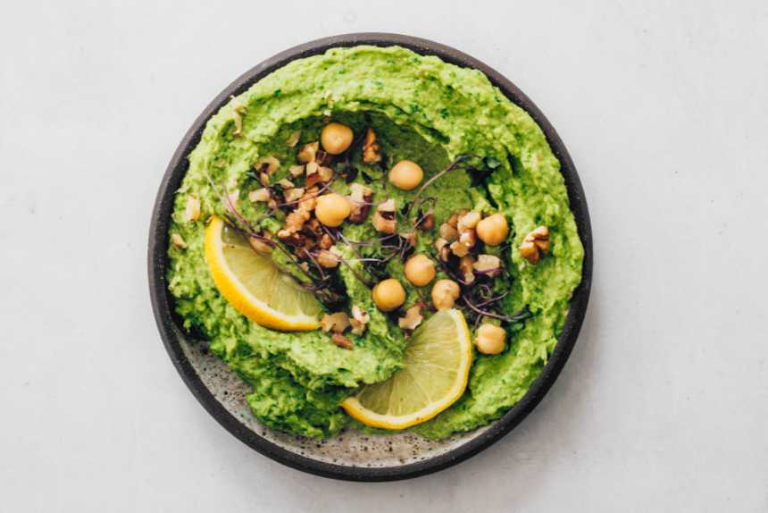 small bowl with green hummus that's topped with chickpeas, walnuts, herbs and lemon slices