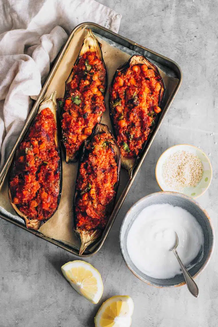 baking dish with four eggplant halves filled with red seasoned quinoa mixture next to some vegan yogurt