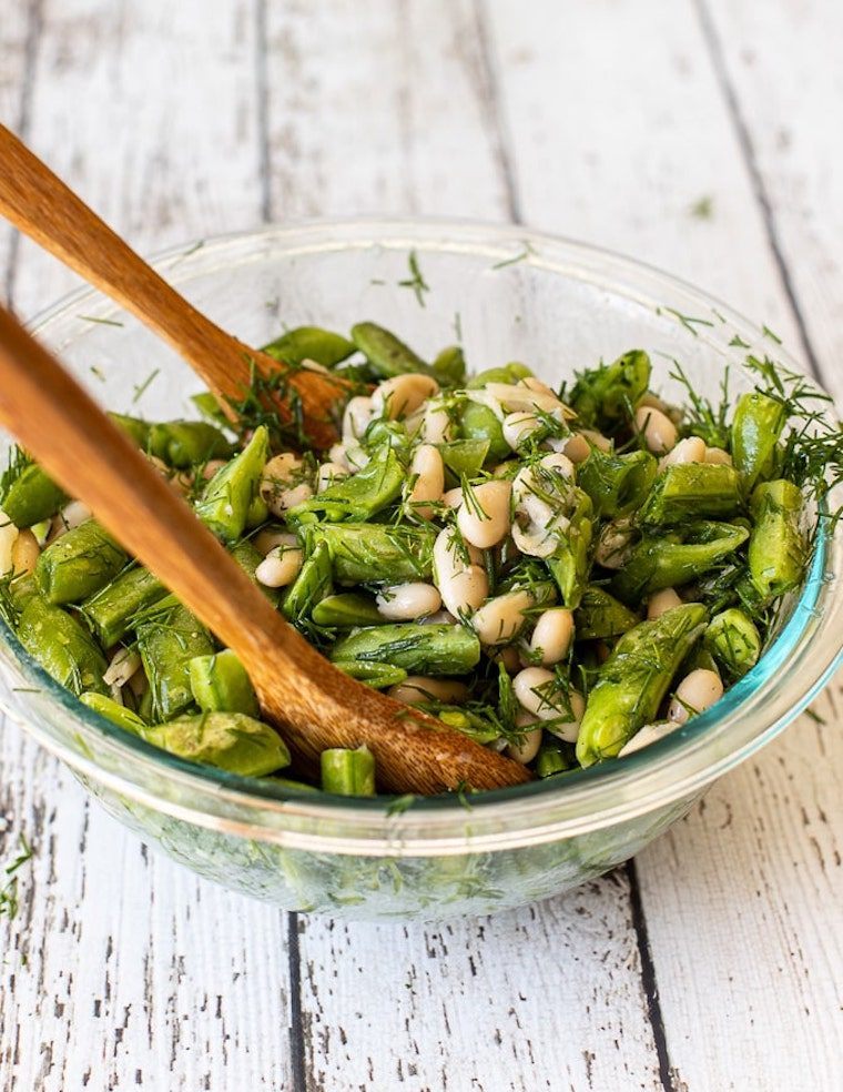 glass bowl on wooden table with green sugar snap peas, white beans and fresh dill