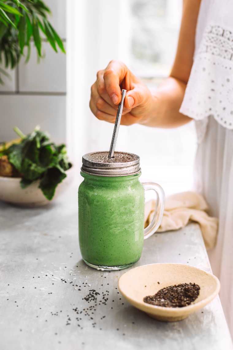 woman in white dress standing by a window and putting a straw in a glass of super green smoothie