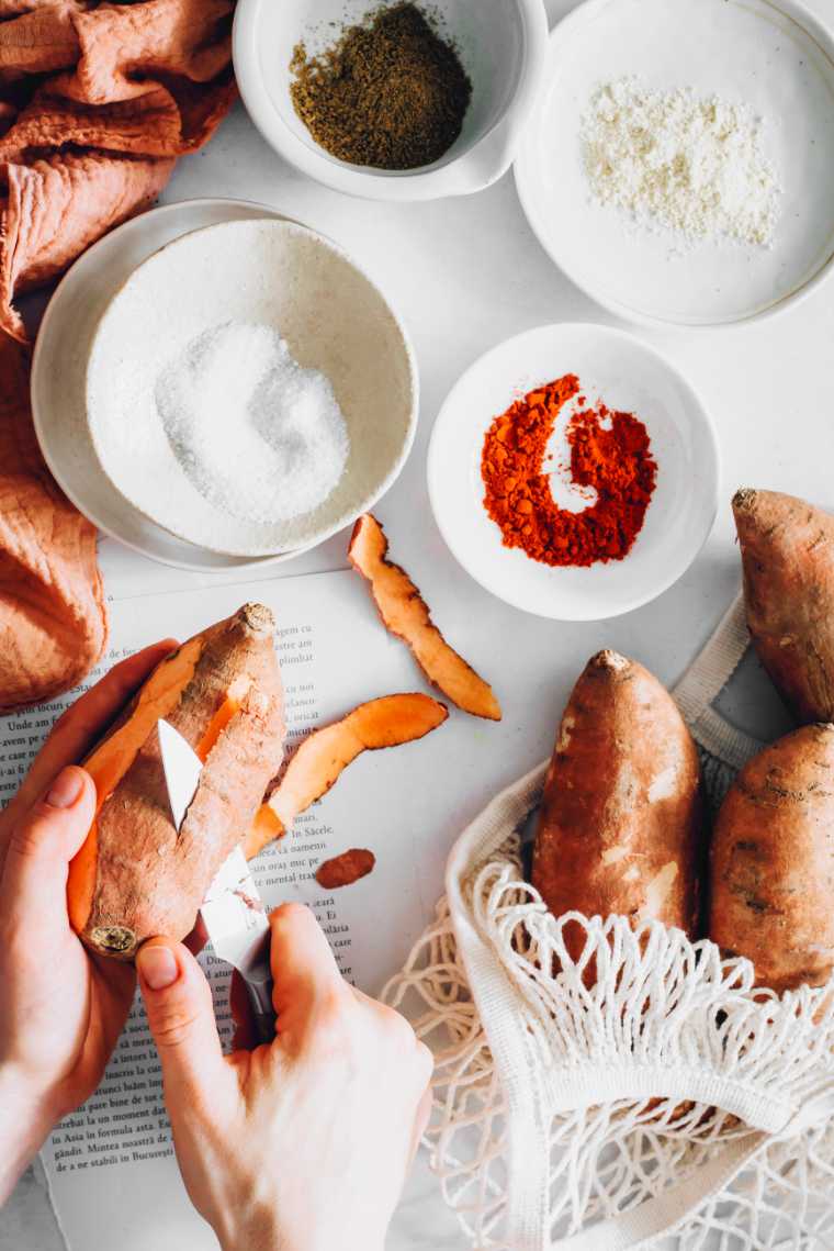 witte tafel met verschillende witte kommen met kleurrijke kruiden erin en twee handen die een zoete aardappel schillen