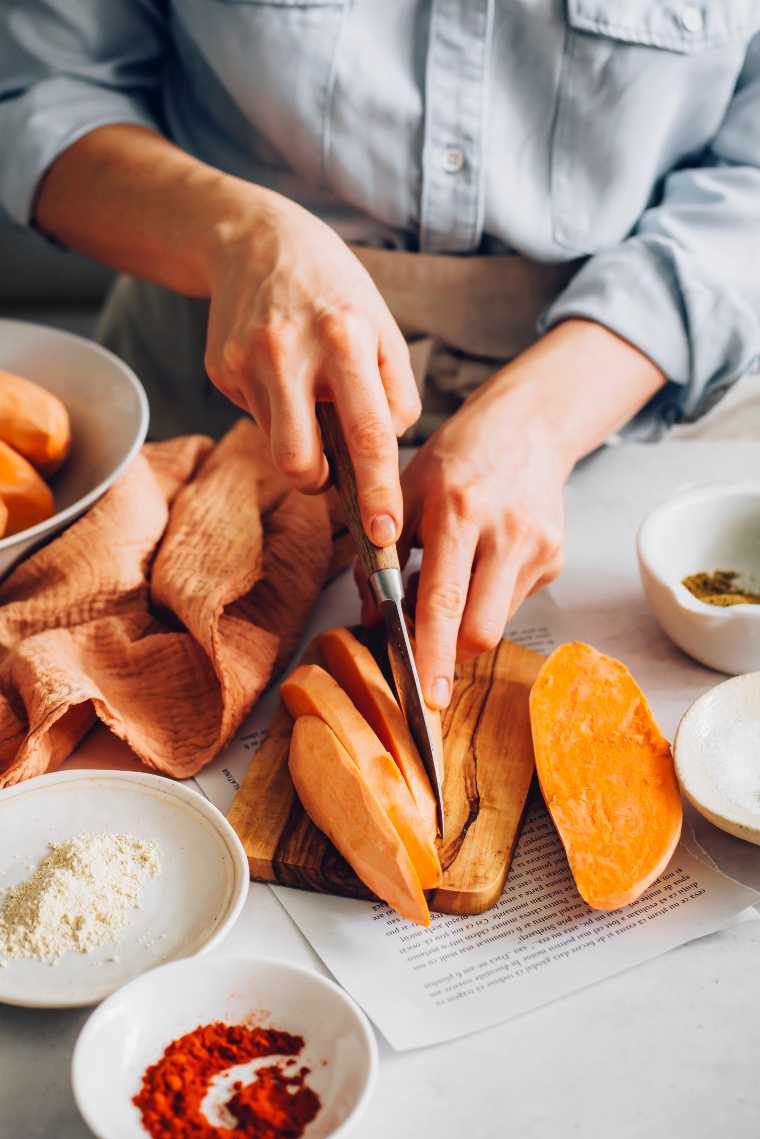 vrouw in blauw overhemd zit aan een witte tafel en snijdt oranje zoete aardappel in plakjes