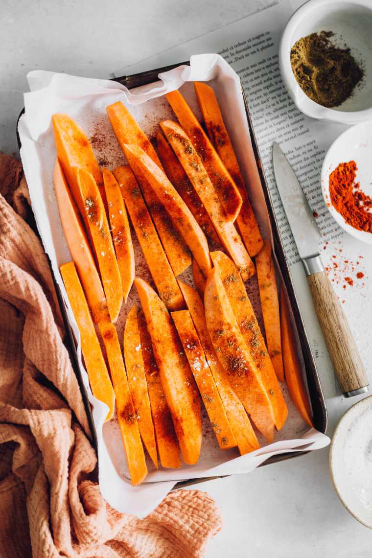 Ein weißer Tisch mit einer mit Pergamentpapier ausgelegten Auflaufform, in der sich jede Menge in Scheiben geschnittene und gewürzte orangefarbene Süßkartoffeln befinden